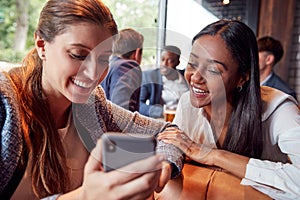 Two Businesswomen Looking At Photos On Mobile Phone In Bar After Work With Colleagues