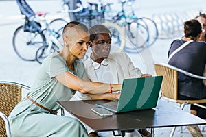 Two businesswomen on the terrace of a coffee shop are discussing a job on the laptop and have a smartphone on the table.