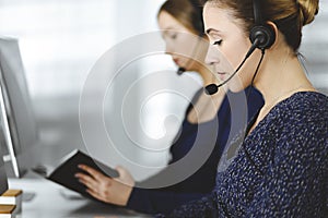 Two busineswomen have conversations with the clients by headsets, while sitting at the desk in a modern office. Diverse