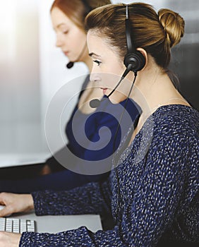 Two busineswomen have conversations with the clients by headsets, while sitting at the desk in a sunny modern office