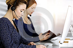 Two busineswomen have conversations with the clients by headsets, while sitting at the desk in a sunny modern office