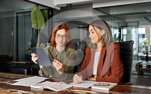 Two busy business women talking using tab working together at desk in office.