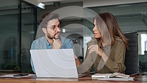 Two busy colleagues working together talking using laptop in office.