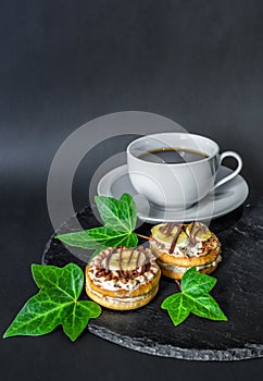 Two cake biscuits with cream, banana and chocolate and a cup of coffee on a slate dish on a black background, decorated with green