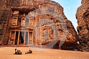 Two camels, saddles ready for tourists, sitting in front of Al-Khazneh Treasury temple - main attraction in Lost city of Petra.