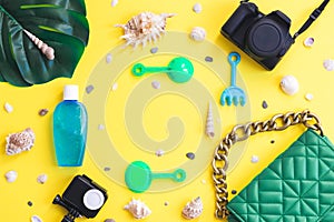 Two cameras, a green bag, a monstera leaf, a children set for sand, seashells and suntan lotion on a yellow background