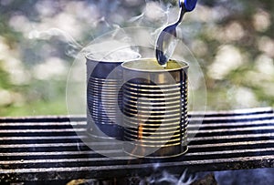 Two cans of food being cooked over a campfire, steam rising, spoon held