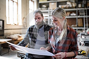 Two carpenters man and woman looking at blueprints indoors in carpentery workshop.
