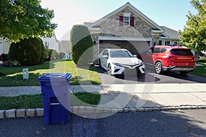Two cars on a asphalt driveway in front of an open garage of a small house with a blue recycle bin near the curb