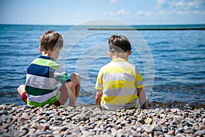 Two Caucasian baby boys is sitting at the beach. Kids in pants with his back sitting on the sea on a rocks pebbles. Sea vacations
