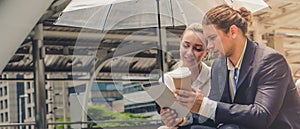 Two caucasian business people holding umbrella sitting outdoor watching tablet computer with coffee cup on hand