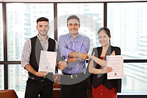 Two caucasian businessmen and asian businesswoman shaking hands while holding success business certificate with happy and smiling