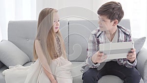 Two Caucasian children sitting on couch. Cute Caucasian girl touching boy`s shoulder and waving. Boy making displeased