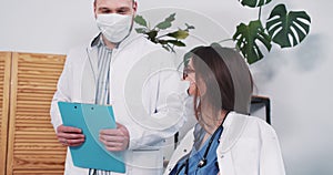 Two Caucasian doctors in lab coats, masks consulting each other at work, discuss case at light clinic office table.