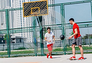 Two Caucasian friends wearing red sports equipment playing basketballs outdoors on a sunny day