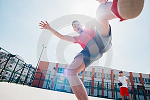 Two Caucasian friends wearing red sports equipment playing basketballs outdoors on a sunny day while they dribble and push each