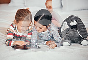 Two caucasian girls using a digital tablet while lying together on a bed at home. Happy young children browsing the