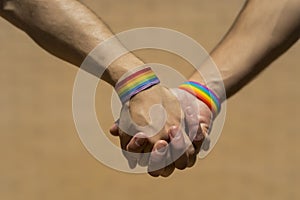 Two Caucasian men holding hands with a rainbow-patterned wristband against a brown background