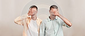 Two caucasian men, twin brothers covering eyes with hand while posing together isolated over beige background, panoramic