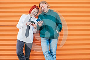 Two caucasian teen friends boy and a girl browsing their smartphone devices while they lean on the orange wall background.