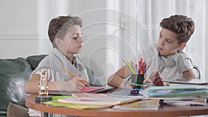 Two Caucasian twin brothers sitting at the table and studying. One boy trying to cheat, his sibling closing his exercise