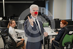 Two Caucasian women look at a serious elderly man with a clown nose in the office.