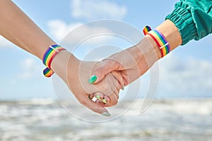 Two caucasian womens holding hands with a rainbow-patterned wristban on their wrists.
