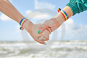 Two caucasian womens holding hands with a rainbow-patterned wristban on their wrists.