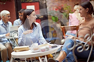 Two caucasian young adult women sitting in the outdoor cafe, talking, socializing, smiling, looking each other