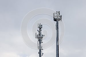 Two cell towers with different antennas and signal boosters against a cloudy sky