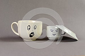 Two ceramic cups against a gray background. A large cup with a drawn surprised woman`s face and puffy lips. A small cup with a ma