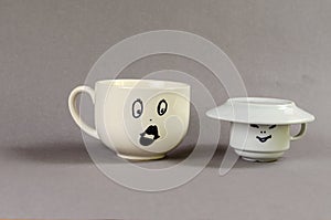 Two ceramic cups against gray background. Large cup with a drawn surprised woman`s face and puffy lips. Small cup with a man`s