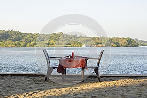 A two chairs and a table with a red tablecloth against the background of a river and green forest. open air restaurant