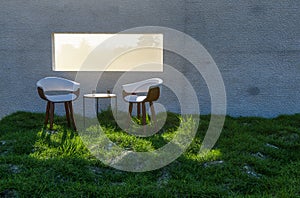 Two Chairs, Table and Water Glasses Near Window with Bright Sun Light Shining Through Concrete Wall in Grass and Rocks Field