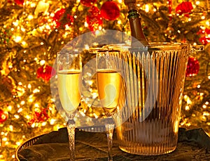 Two champagne flutes and a champagne bottle in a glass ice bucket in front of a Christmas tree.