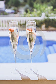 Two champagne glasses with strawberry on sunny terrace outdoor patio overlooking swimming pool at summer day outside of