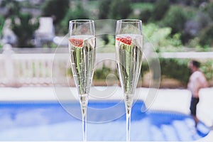 Two champagne glasses with strawberry on sunny terrace outdoor patio overlooking swimming pool at summer day outside of