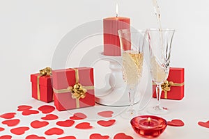 Two champagne glasses on a white background. Decorated with burning candles, red hearts, red boxes with gifts. Champagne