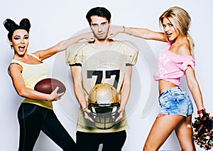 Two charming cheerleaders with a ball are striking on a quarterback American football player with a helmet.