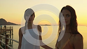 Two charming female models in sundresses pose on promenade by the ocean on beautiful summer evening