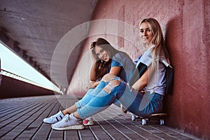 Two charming hipster girls sitting on skateboard and leaning on a wall at the sidewalk.
