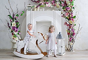 Two charming little girls play in the light room decorated with flowers. Baby girl swinging on a wooden horse
