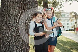 Two charming school-age girls stand by the tree near the school and play on the tablet