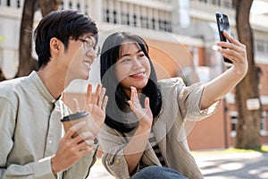 Two cheerful Asian friends are taking pictures together with a phone while chilling in a city park