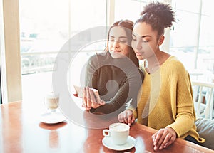 Two cheerful and beautiful girls are sitting together near the table and watching something on the phone. They look