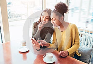 Two cheerful and beautiful girls are sitting together near the table and watching something on the phone. They look