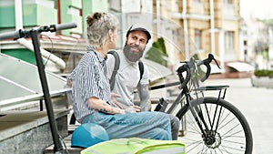 Two cheerful couriers, young man and woman sitting on the bench and talking outdoors while delivering food and products