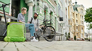 Two cheerful couriers, young man and woman sitting on the bench and talking outdoors while delivering food and products