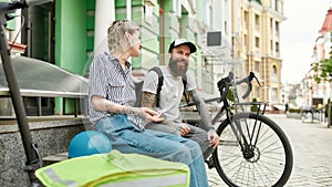 Two cheerful couriers, young man and woman sitting on the bench and talking outdoors while delivering food and products