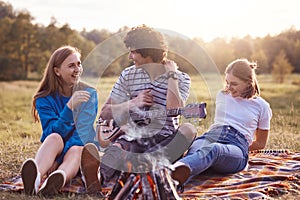 Two cheerful cute girls spend free time with handsome curly male who plays guitar and entertains them, pose together on plaid near